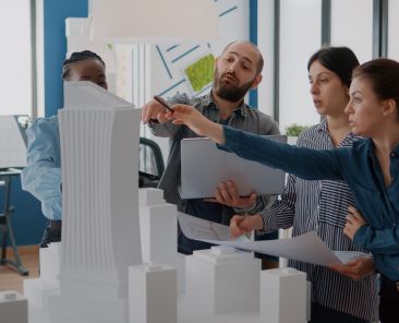 Group of colleagues meeting to analyze building model and maquette to plan blueprints. People working on architecture design with laptop, tablet and urban layout on table. Development