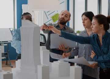 Group of colleagues meeting to analyze building model and maquette to plan blueprints. People working on architecture design with laptop, tablet and urban layout on table. Development